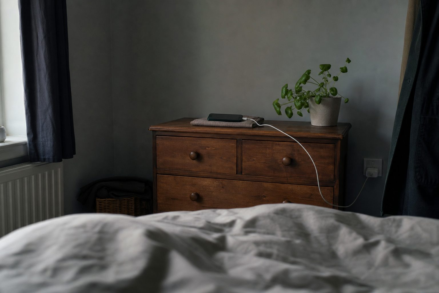 A smartphone charging on a folded cloth on top of a wooden chest of drawers across a dim bedroom, seen from the bed